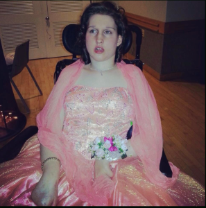 Stephanie is sitting in her wheel chair in a pink prom dress with a corsage of white and pink flowers around her left wrist. she is looking to the right