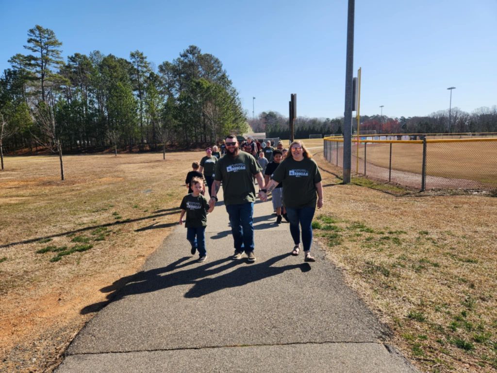Dawson on the left then his dad then his mom are walking on the paved path with grass on both sides. They are walking towards the camera with the other walkers behind them