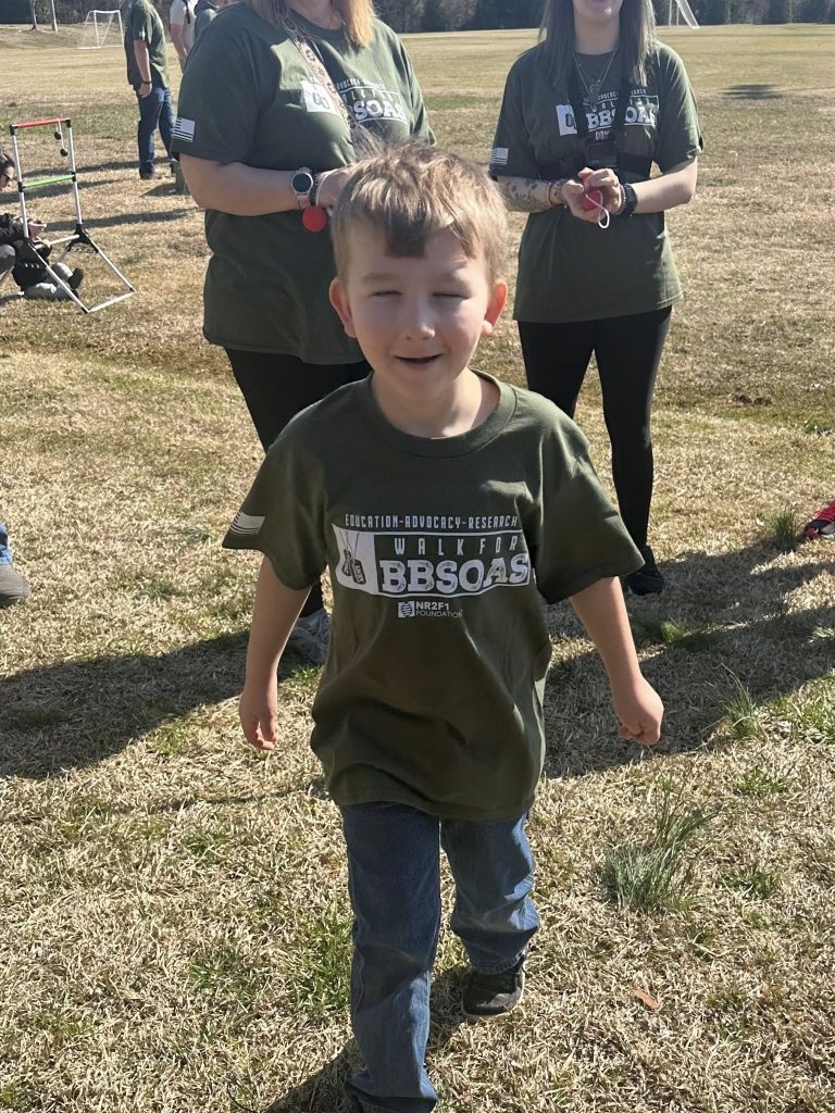 Dawson is walking on grass towards the camera wearing a green t-shirt that says walk for BBSOAS. He is smiling at the camera. There are two participants standing behind him.