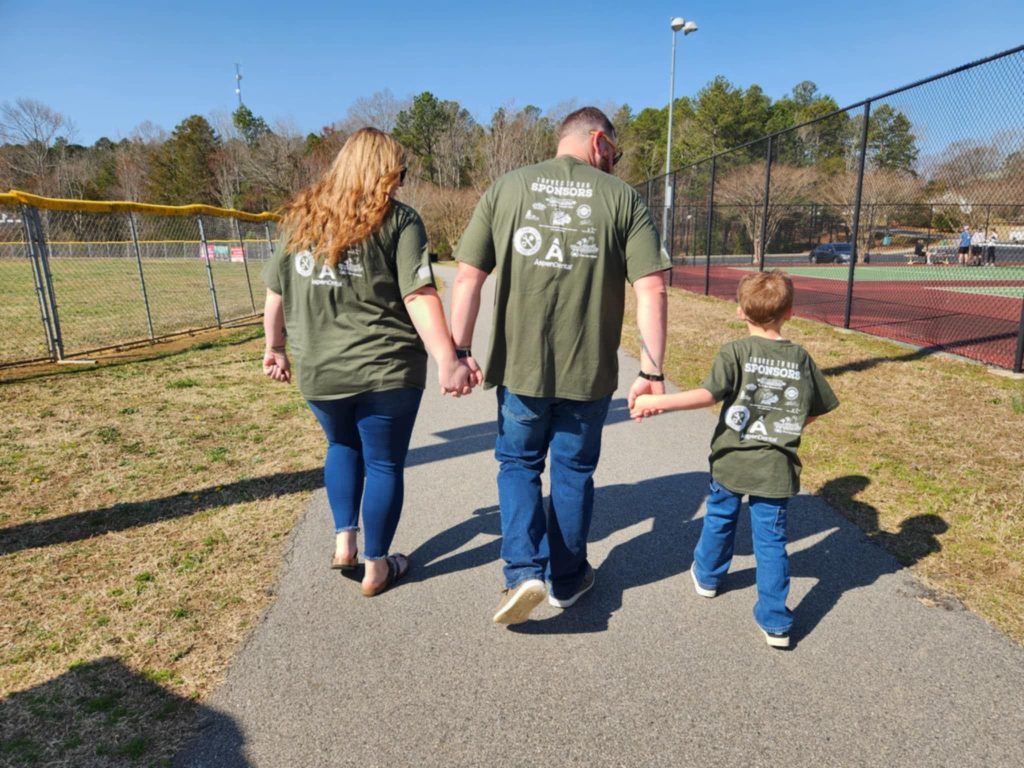 from left to right we see the backs of Dawson's mom holding Dawson's dad's hand to her right and dad is holding Dawson's hand as they walk a paved path at the park. They are wearing green t-shirts with business logos on the back. They are all wearing blue jeans.
