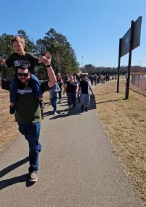 Dawson is sitting on the shoulders of his father and holding his hands. they are walking down a paved path with other walkers behind them